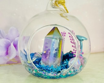 May include: A clear glass terrarium with a large, iridescent crystal at its center. The terrarium is filled with blue and purple decorative elements, including moss and small decorative pieces. A small, blue mushroom and purple plant are also inside.