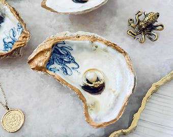 May include: Decorative oyster shell ring dishes with gold trim and blue and white designs. A gold octopus figurine and a gold pendant necklace are also present. One shell holds a ring with a pearl.