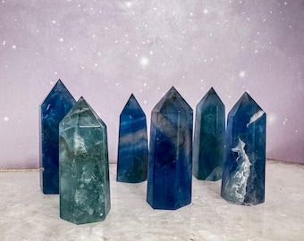 May include: A collection of six polished fluorite crystal points in varying shades of deep blue, teal, and green. Each stone features a hexagonal shape with a pointed top, displaying natural internal banding, cloudy inclusions, and translucent areas.
