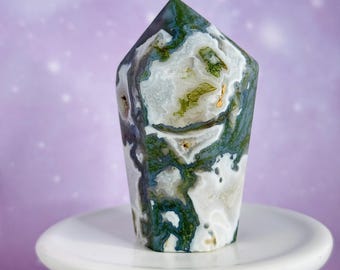 May include: A polished gemstone obelisk with a flat top. The stone features a mix of white, green, and blue-grey patterns. It sits on a small, white, round pedestal against a soft purple background.