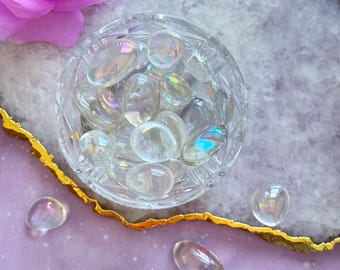 May include: A clear, cut-glass bowl filled with iridescent, oval-shaped stones. Several stones are scattered around the bowl on a purple surface with a gold edge. A large, pink rose is in the upper left corner.