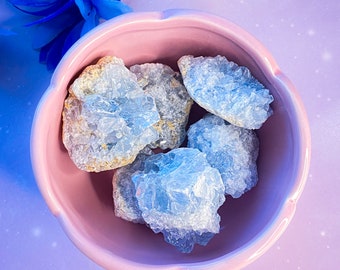 May include: A pink, scalloped-edge bowl filled with light blue celestite crystals. The crystals have a rough, textured appearance. Blue artificial flowers are in the background, set against a purple and white speckled background.