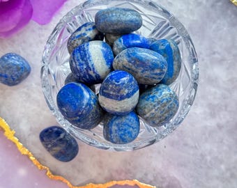 May include: A clear glass bowl filled with polished lapis lazuli stones. The stones are a deep blue color with white and gray marbling. Some stones are scattered around the bowl on a purple surface with a gold trim.