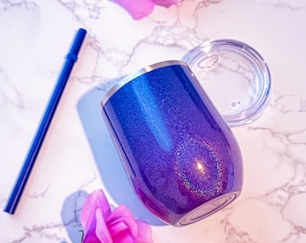May include: A blue and purple glitter tumbler with a clear lid and a blue straw. The tumbler has a silver rim and is next to two pink roses on a marble surface. The tumbler is designed to keep drinks cold.