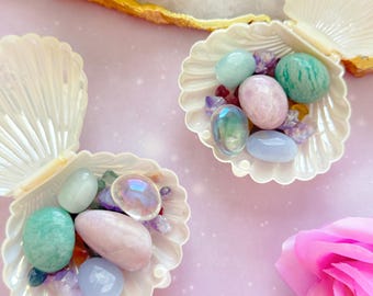 May include: Two open, iridescent white seashell bowls filled with colorful gemstones and crystals. The gemstones are various shapes and sizes, including pink, blue, green, and clear. A pink flower is in the lower right corner, and the background is a light purple.