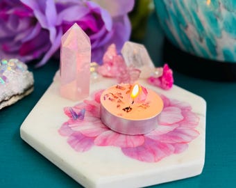May include: A lit candle in a silver holder sits on a white hexagonal marble tray decorated with a pink floral design. A pink crystal and other crystals are arranged around the candle. Purple flowers are in the background.