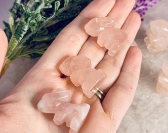 May include: A hand holds several small, carved rose quartz bunny figurines. The bunnies are a soft pink color and have detailed features. Purple flowers and greenery are in the background, adding a touch of color.