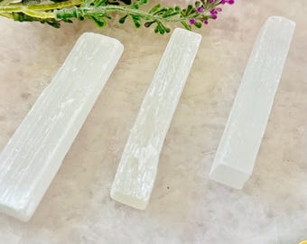 May include: Three white selenite crystal wands are arranged on a decorative surface, with a sprig of purple flowers in the background. The wands have a smooth, elongated shape and a translucent appearance. The crystals are a popular choice for spiritual practices.