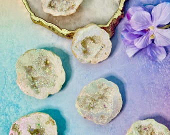 May include: Six iridescent geode slices with crystal formations. The geodes are displayed on a white and gold-edged surface, with a gradient blue and purple background and a purple flower.
