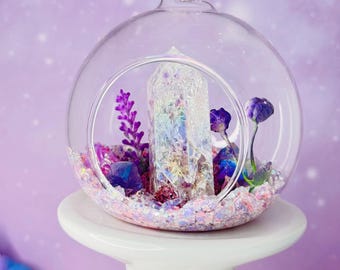 May include: A clear glass terrarium with a central crystal, surrounded by purple and pink decorative elements. The terrarium is suspended by a gold-colored cord and sits on a white pedestal, with a soft purple background.
