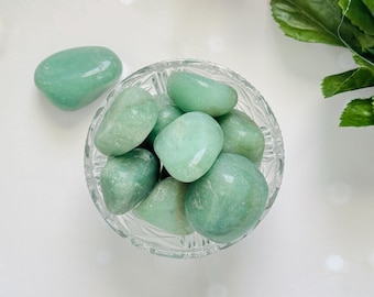 May include: A top-down view of a clear glass bowl filled with smooth, polished, light green stones. One stone rests outside the bowl. Purple and blue flowers are in the background.