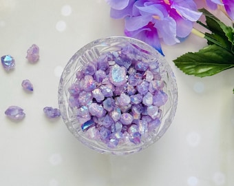 May include: A clear, cut-glass bowl filled with iridescent purple crystals. Several loose crystals are scattered around the bowl. Artificial purple flowers are in the background, creating a decorative display. The crystals have a shimmering, multi-colored effect.