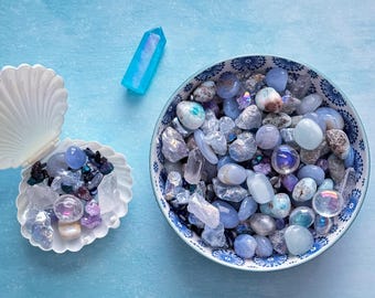 May include: A collection of polished gemstones and crystals in shades of blue, purple, and clear. The stones are displayed in a seashell and a decorative bowl with a blue and white pattern. A blue crystal wand is also present.