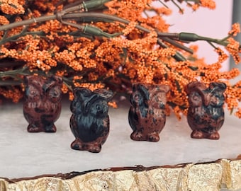 May include: Four small owl figurines in shades of brown and black are arranged on a white surface. The owls have detailed features and a textured appearance. Orange flowers are in the background.