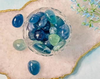 May include: A clear glass bowl filled with polished blue and green gemstones. Several stones are scattered around the bowl on a white and pink tray with a gold edge. Blue flowers are in the background.