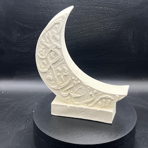 May include: A cream-coloured crescent moon sculpture with intricate carved Arabic calligraphy. The moon sits atop a rectangular base, displayed on a black rotating stand. The sculpture is a decorative piece, likely for home decor.