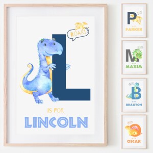 May include: Watercolor dinosaur art print featuring a blue dinosaur with a yellow belly and the letter "L" for "Lincoln". The print includes the text "ROAR!" in a speech bubble and is framed in a light wood frame. Additional prints with other names are also shown.