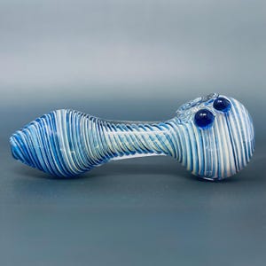 Könnte beinhalten: Eine Glaspfeife mit blau-weißem Streifendesign. Die Pfeife hat einen abgerundeten Kopf mit zwei dunkelblauen Glasakzenten und einen langen, zylindrischen Körper. Das Mundstück ist aus klarem Glas.