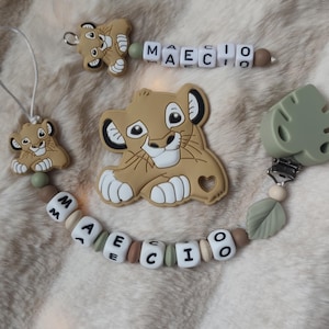 Peut inclure: Ensemble d'accessoires pour bébé sur le thème du lionceau. L'ensemble comprend un attache-tétine, une tétine et un anneau de dentition. Le clip et l'anneau de dentition ont le nom "MAECIO" épelé en perles de lettres blanches. Le lionceau est beige avec un visage blanc.