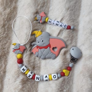Peut inclure: Ensemble de clips et de breloques pour bébé avec un éléphant gris aux oreilles roses et un chapeau jaune. Le clip et le charme portent le nom "HINAO" épelé en perles de lettres blanches. L'ensemble comprend un clip de sucette, un charme et une plus grande figurine d'éléphant.