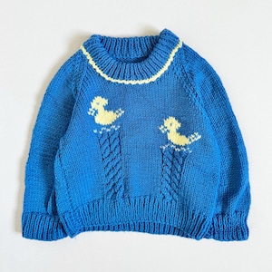 May include: A blue knitted sweater with yellow duck designs. The sweater features a ribbed collar with yellow trim and cable knit detailing. The ducks are yellow and appear to be floating on water. The sweater is a children's size.