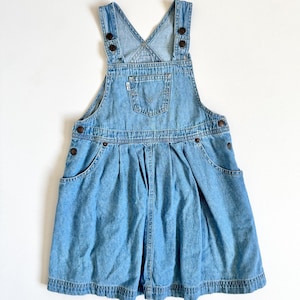 Puede incluir: Una taza de cerámica blanca con asa negra y el texto "But First, Coffee" en negrita negra. La taza se encuentra sobre un fondo borroso, sugiriendo un ambiente acogedor.