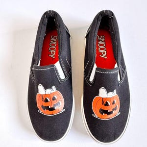 May include: Pair of black slip-on shoes featuring a Snoopy-themed jack-o'-lantern design. The shoes have white soles and trim, with the word "SNOOPY" printed on the red insole. A fun Halloween-themed footwear option.