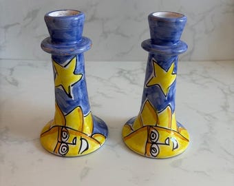 May include: A pair of ceramic candle holders featuring a hand-painted blue background with yellow stars and stylised yellow sun faces at the base. Each holder has a flared bottom and a cylindrical top section designed to hold a taper candle.