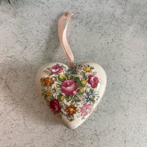 May include: A heart-shaped ceramic ornament featuring a cream-colored base decorated with a colorful floral pattern of pink, red, yellow, and blue flowers with green leaves. A light pink ribbon is attached to the top for hanging purposes.