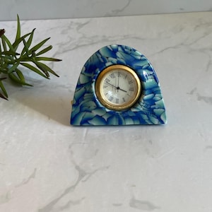 May include: A small, arched clock with a blue and white marbled design. The clock face is gold with black Roman numerals and black hands. The clock is a decorative item.
