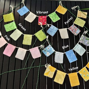 Op de afbeelding: Een kleurrijk stoffen banner met vlaggen in tinten roze, groen, blauw, geel en rood. De vlaggen zijn aan elkaar geregen met groen draad en hebben verschillende patronen. De banner is gelabeld met de woorden "Roze", "Groen", "Vibrant", "Pastel", "Blauw" en "Geel".