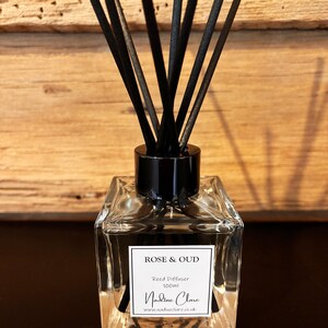 May include: A clear, square glass reed diffuser with black reeds. The label reads "ROSE & OUD, Reed Diffuser 100ml, Nadine Clare, www.nadineclare.co.uk". The diffuser sits on a dark surface against a wooden background.