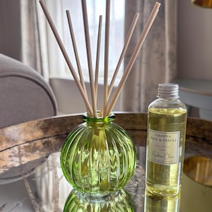 May include: A green ribbed glass reed diffuser bottle with several light-coloured reeds inserted, accompanied by a clear plastic refill bottle containing yellow liquid with a label reading Verbena Basil & Neroli Reed Diffuser 100ml Nadine Clare.