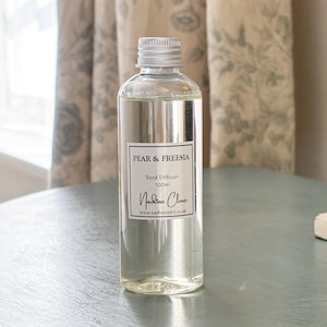 May include: A clear glass bottle of reed diffuser oil with a silver screw cap. The white label features the text PEAR & FREESIA, Reed Diffuser, 100ml, Nadine Clare, and the website address www.nadineclare.co.uk in black lettering.