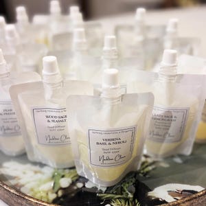 May include: Several clear pouches filled with liquid, each with a white nozzle and a label. The labels feature text like "Wood Sage & Seasalt" and "Verbena Basil & Neroli". These pouches are refills for reed diffusers.