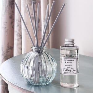 May include: A clear glass reed diffuser with grey reeds and a matching glass bottle of Wood Sage & Seasalt fragrance. The diffuser has a ribbed design and sits next to a 100ml bottle with a label that reads "Nadine Clare."