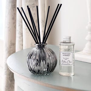 May include: A stylish reed diffuser set featuring a ribbed glass vase with black reeds. A clear bottle of Wood Sage & Seasalt Reed Diffuser, 100ml, sits beside the vase. The set is on a light blue surface.