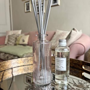 May include: A clear, vertically ribbed glass reed diffuser bottle containing several grey reeds, accompanied by a separate clear glass refill bottle with a silver cap and a white label reading Wood Sage & Seasalt Reed Diffuser 100ml Nadine Clare.