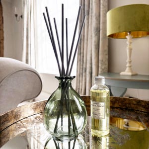 May include: A green glass diffuser bottle with a narrow neck containing several dark grey reed sticks, accompanied by a clear plastic refill bottle labelled Verbena Basil & Neroli with a silver cap, containing 100ml of scented liquid fragrance.