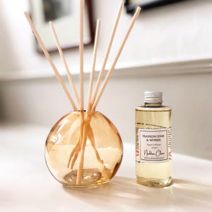 May include: An amber glass reed diffuser with wooden reeds, and a small bottle of Frankincense & Myrrh Reed Diffuser. The bottle has a silver cap and a label that reads "100ml". The liquid inside the bottle is light yellow.