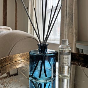 May include: A blue glass reed diffuser bottle with several black reeds inserted, accompanied by a clear plastic refill bottle labelled Wood Sage & Seasalt Reed Diffuser 100ml by Nadine Claire, featuring a silver screw cap and clear liquid contents.