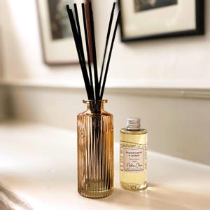 May include: A gold-toned glass reed diffuser with black reeds and a small bottle of essential oil. The diffuser bottle has vertical ridges. The oil bottle has a silver cap and a label that reads "Frankincense & Myrrh".