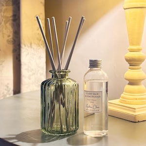 May include: A green glass reed diffuser with white reeds and a clear bottle of liquid. The bottle has a label that reads "Clary Sage & Lemongrass" and "Reed Diffuser 100ml". The diffuser is on a table.