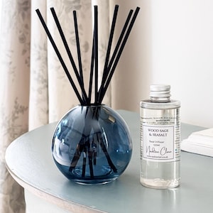 May include: A blue glass reed diffuser with black reeds and a clear bottle of Wood Sage & Seasalt fragrance. The diffuser is round and sits on a light blue surface. The bottle has a white label with text.