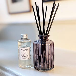 May include: A decorative purple glass reed diffuser with black reeds, alongside a bottle of Wood Sage & Seasalt reed diffuser oil. The oil bottle has a silver cap and a label. A home fragrance item.