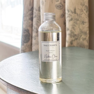May include: A clear glass bottle containing 100ml of reed diffuser oil. The bottle has a silver screw-top lid and a white rectangular label with black text that reads Nag Champa, Reed Diffuser, 100ml, Nadine Clare, and www.nadineclare.co.uk.