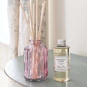 May include: A pink glass reed diffuser with wooden sticks, alongside a 100ml bottle of Frankincense & Myrrh reed diffuser oil. The items are on a light blue surface, with a floral curtain behind.