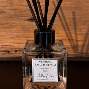 May include: A clear glass reed diffuser with black reeds. The label reads "Verbena Basil & Neroli" and "Reed Diffuser 100ml". The brand name "Nadine Clare" is also visible. The diffuser sits on a dark surface.