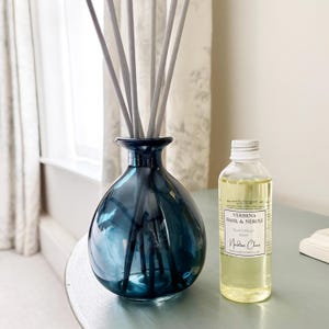 May include: A dark blue glass reed diffuser vessel with several grey reeds inserted, accompanied by a clear plastic bottle containing yellow liquid with a label reading Verbena Basil & Neroli Reed Diffuser 100ml Nadine Clare.