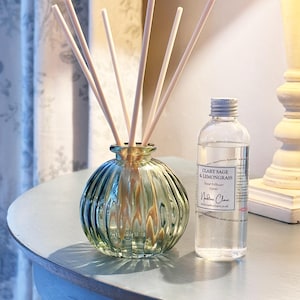 May include: A green glass reed diffuser with wooden sticks and a clear glass bottle. The bottle has a silver cap and a label that reads "Clary Sage & Lemongrass". The diffuser is on a light blue surface.